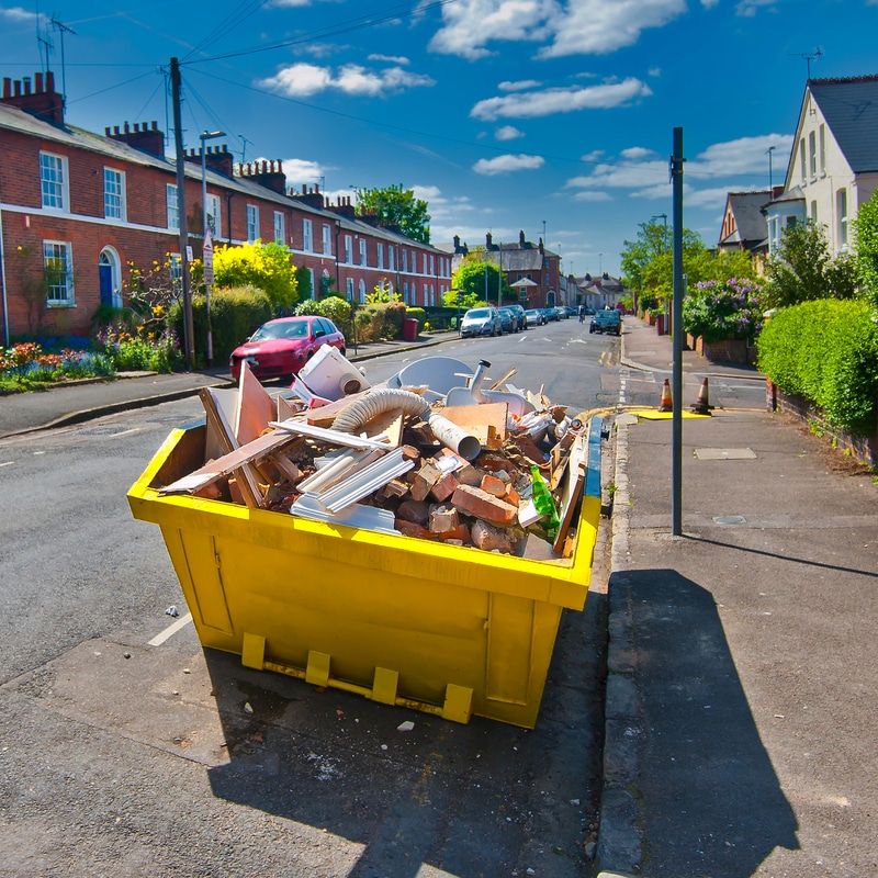 Benne à gravats jaune pour évacuation de déchets de chantier Benne à gravats jaune remplie de débris de chantier sur une rue résidentielle bordée de maisons de briques rouges.